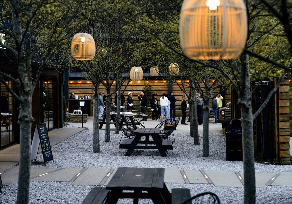 Outdoor courtyard with picnic tables, hanging lanterns, and trees, where a group of people gather and mingle near a wooden building in the evening.