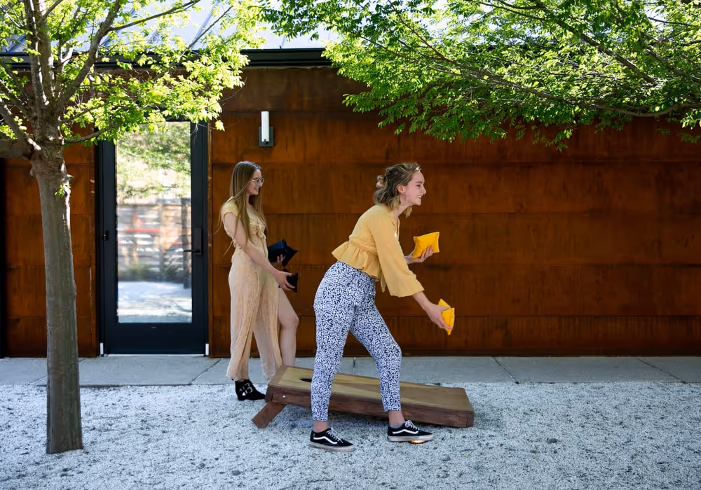 Two women playing an outdoor bean bag toss game on a sunny day, with one throwing a yellow bag while the other watches, standing near a rust-colored wall under leafy green trees.