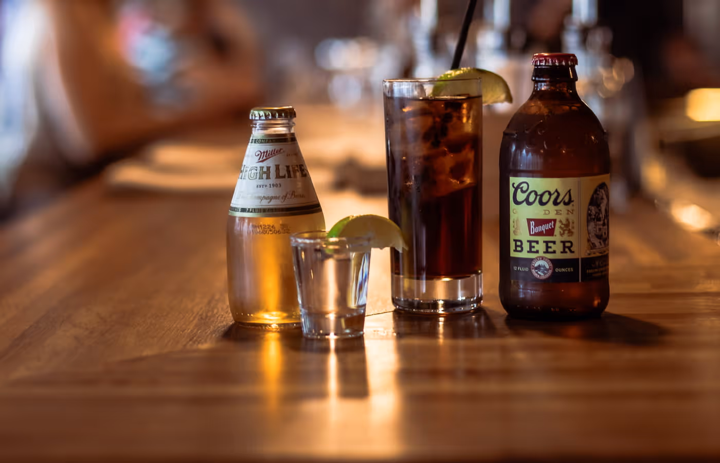 Miller High Life and Coors Banquet beer bottles with shot glass and cocktail with lime on wooden bar counter
