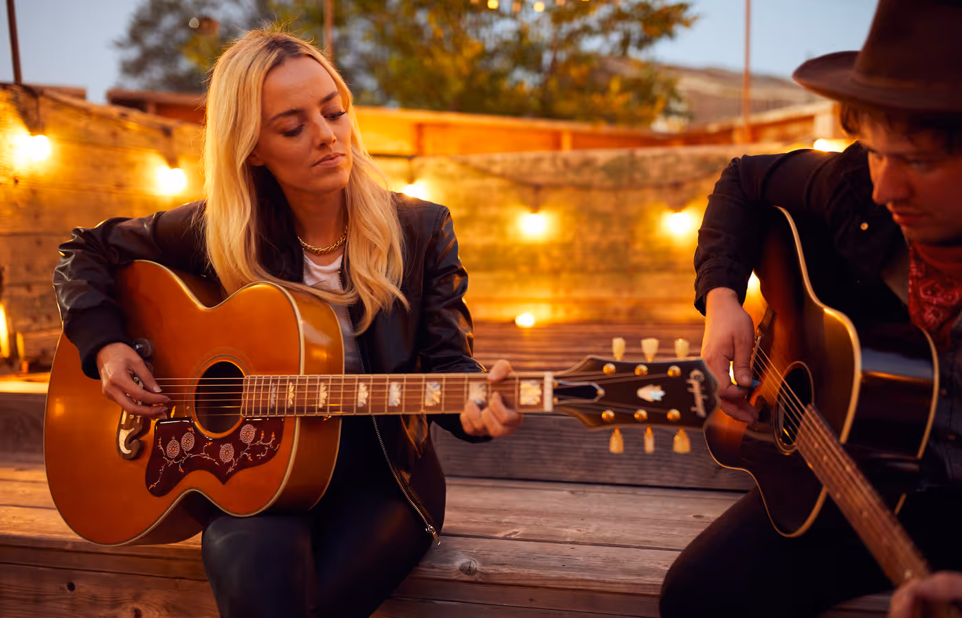 Two musicians playing acoustic guitars outdoors on a wooden deck at dusk with warm string lights in the background.