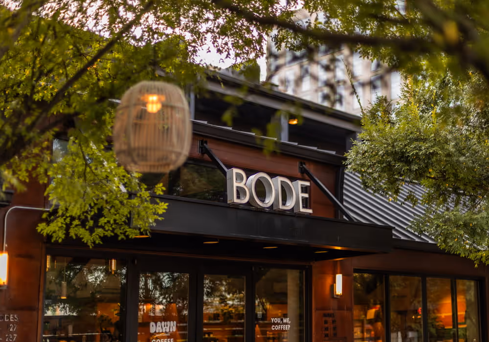 The exterior of Bode hotel with illuminated signage, a wicker lantern, and lush green tree branches framing the view.