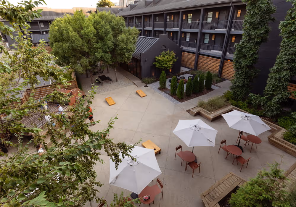Aerial view of a hotel courtyard with white umbrella-shaded patio tables, cornhole boards, and lush greenery