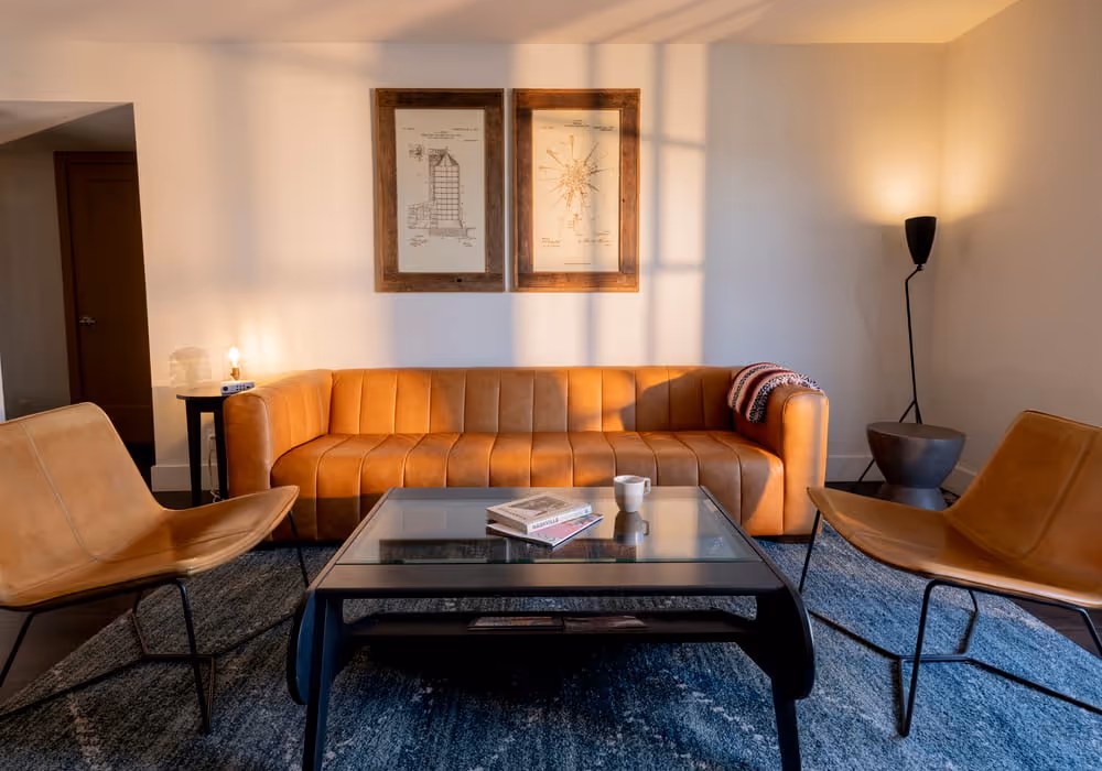 A modern living room featuring a tan leather channel-tufted sofa, two leather accent chairs, and a glass coffee table.