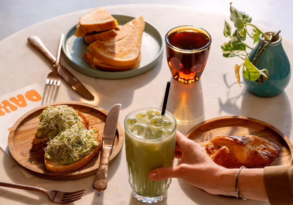 A hand holds an iced matcha latte on a café table with avocado toast, a croissant, sandwiches, and tea at Dava café.