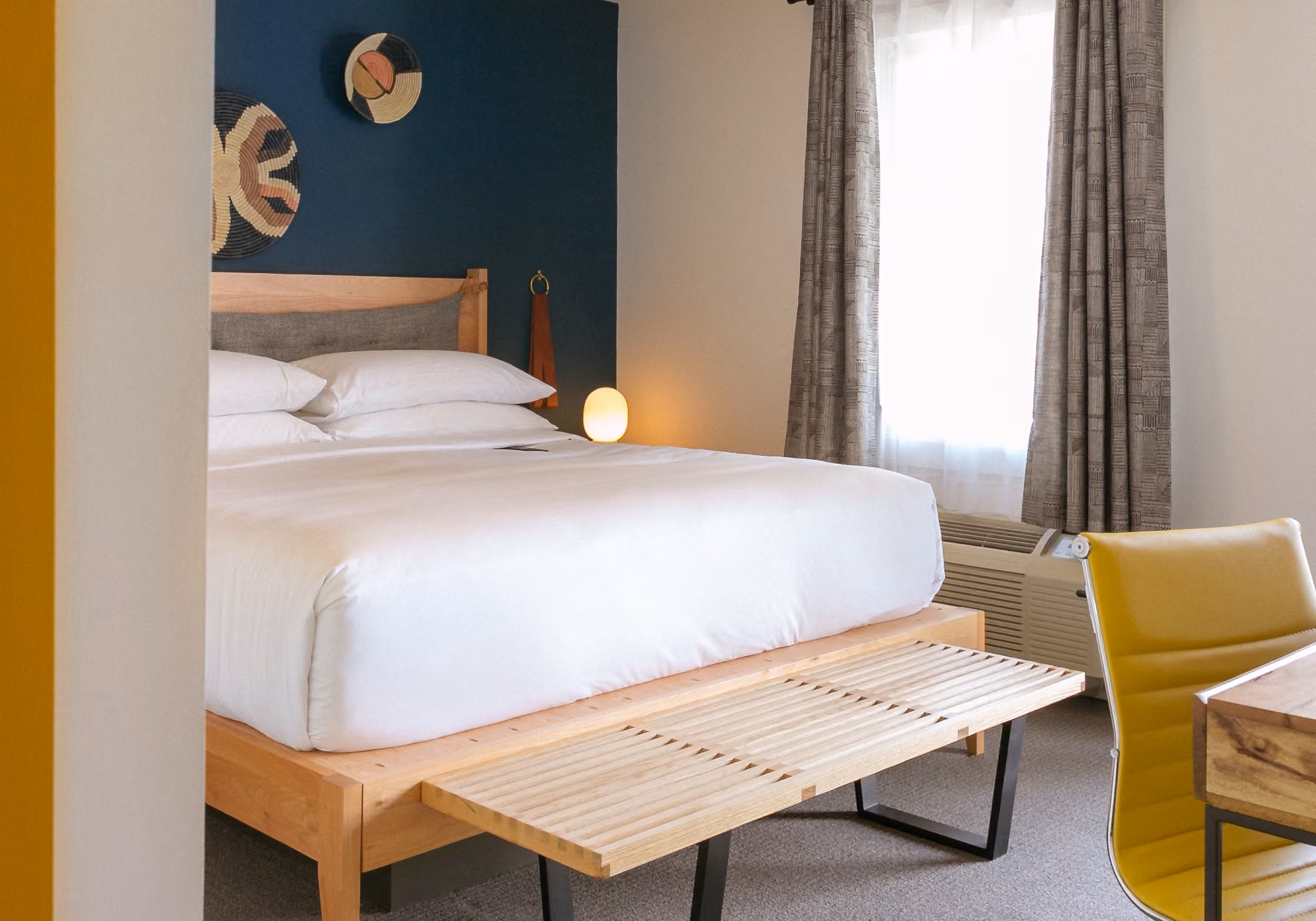 Modern hotel bedroom with white bedding, wooden bed frame, slatted bench, deep teal accent wall, and a yellow leather chair.