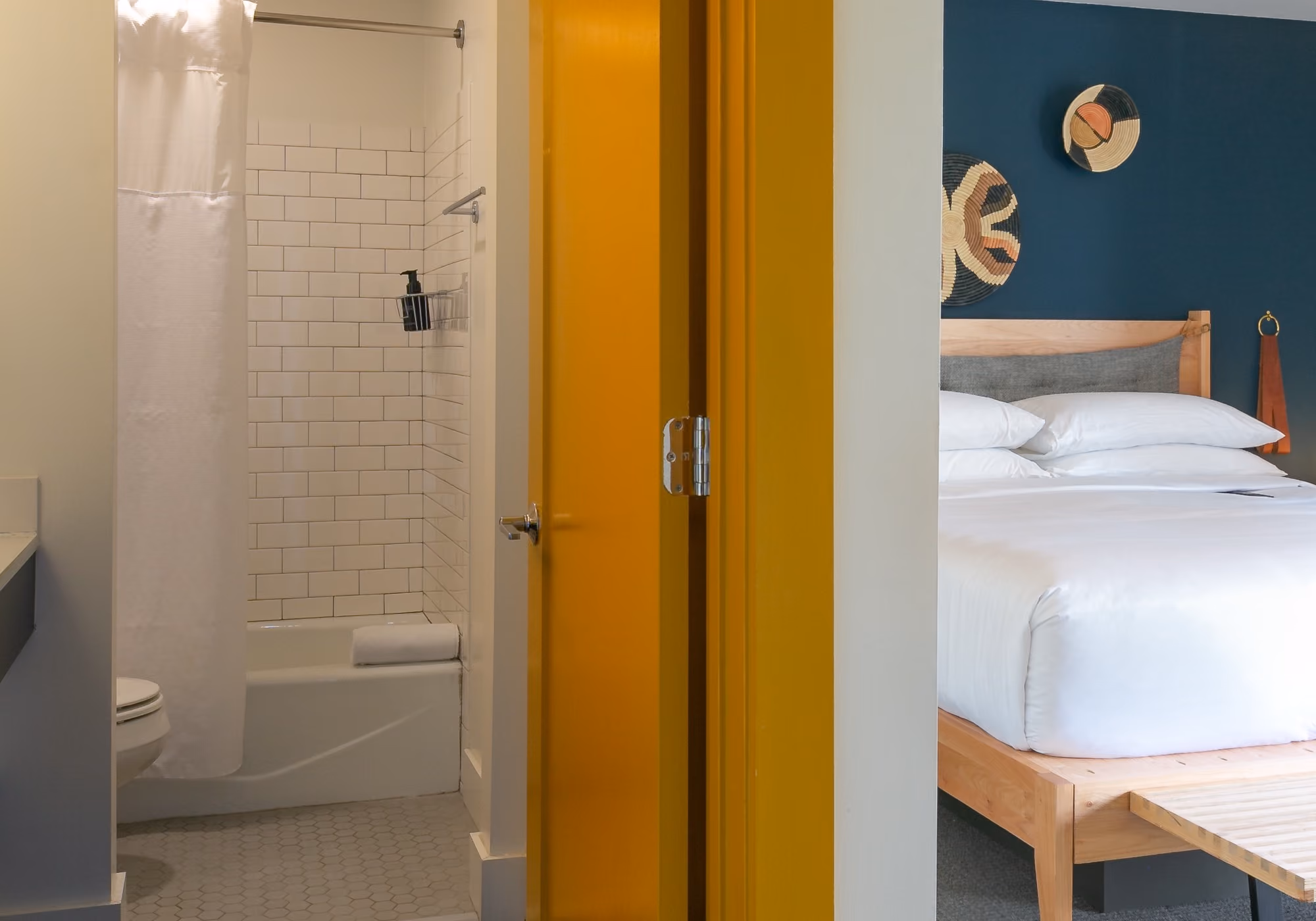 Hotel room view showing white-tiled bathroom with shower, bold mustard yellow door, and bedroom with teal accent wall