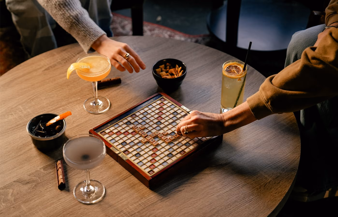 Two people play Scrabble on a round wooden table surrounded by craft cocktails and snack bowls in a cozy bar setting.