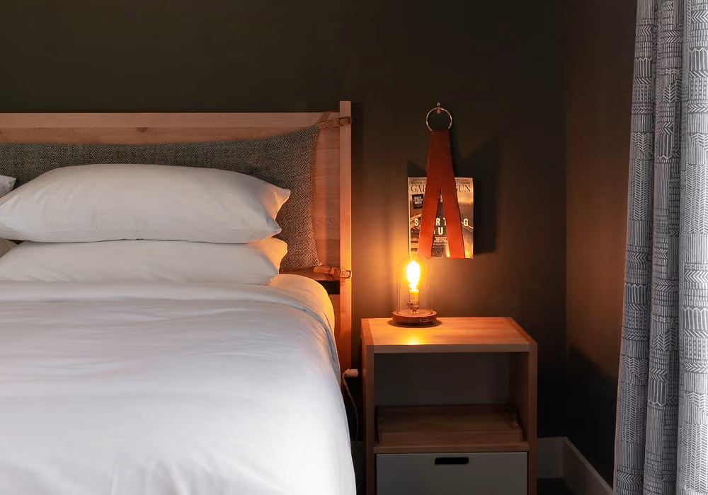 Cozy hotel bedroom featuring crisp white linens, a wooden nightstand with an Edison bulb lamp, and dark green walls