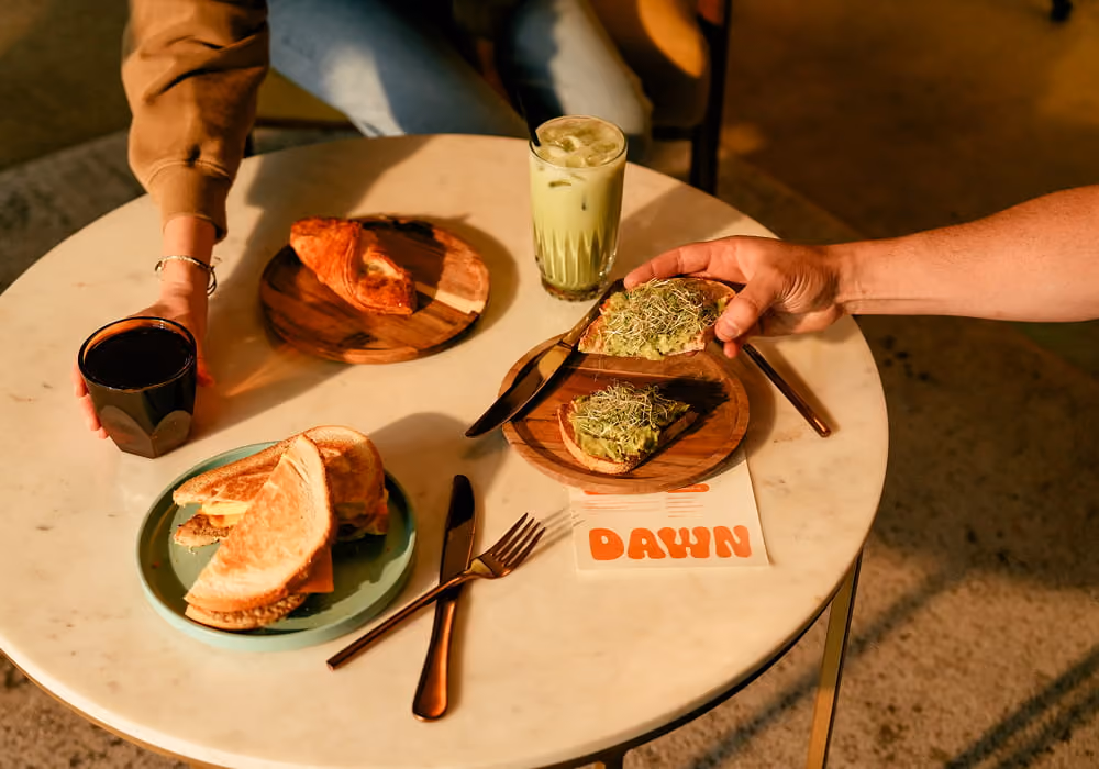 Two guests sharing avocado toast, a croissant, a sandwich, and an iced matcha latte at Dawn café's marble table
