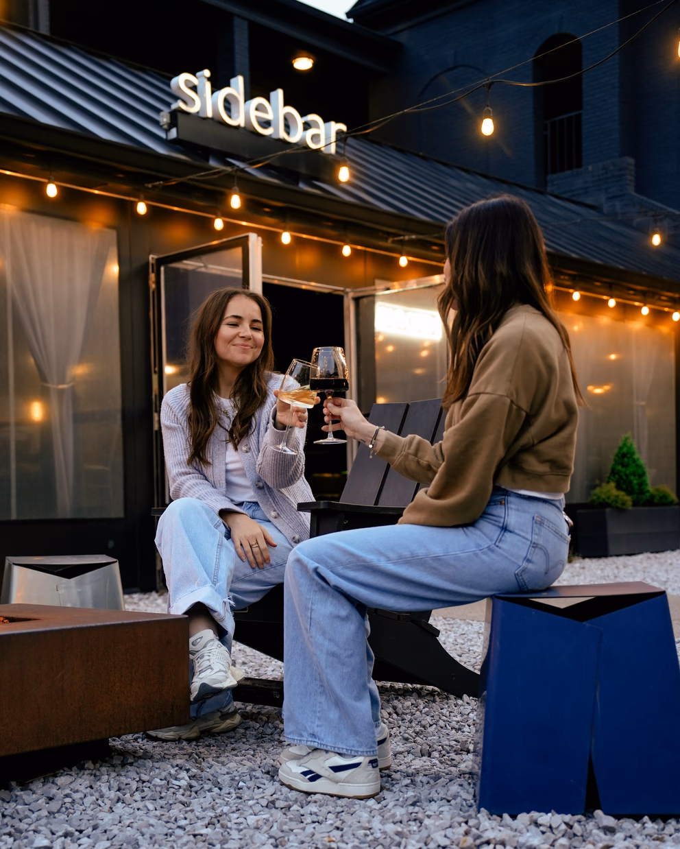 Two friends toasting wine under string lights outside Sidebar bar at Bode Nashville