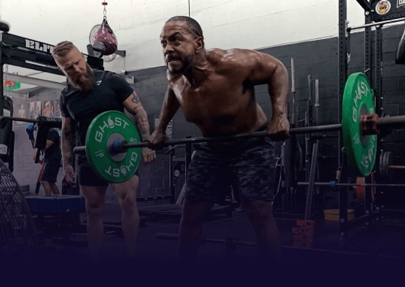 A man is lifting weights in a gym with a trainer standing next to him. The strain is showing on his face and the trainer studies his technique.