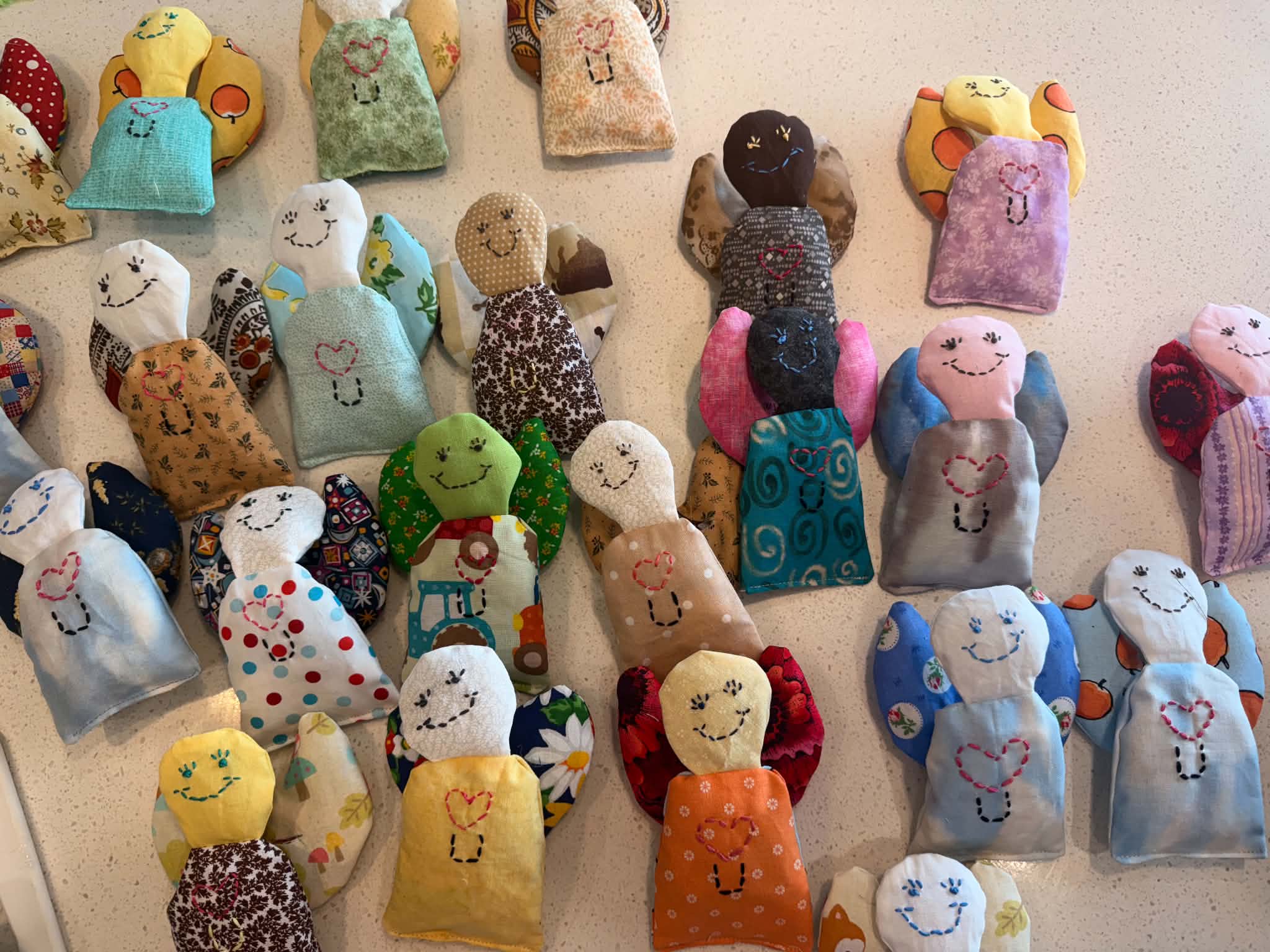 Dozens of handmade Ignite Humanity Angels laid out together in mismatched fabrics, each with a stitched heart, a U, and its own crooked smile