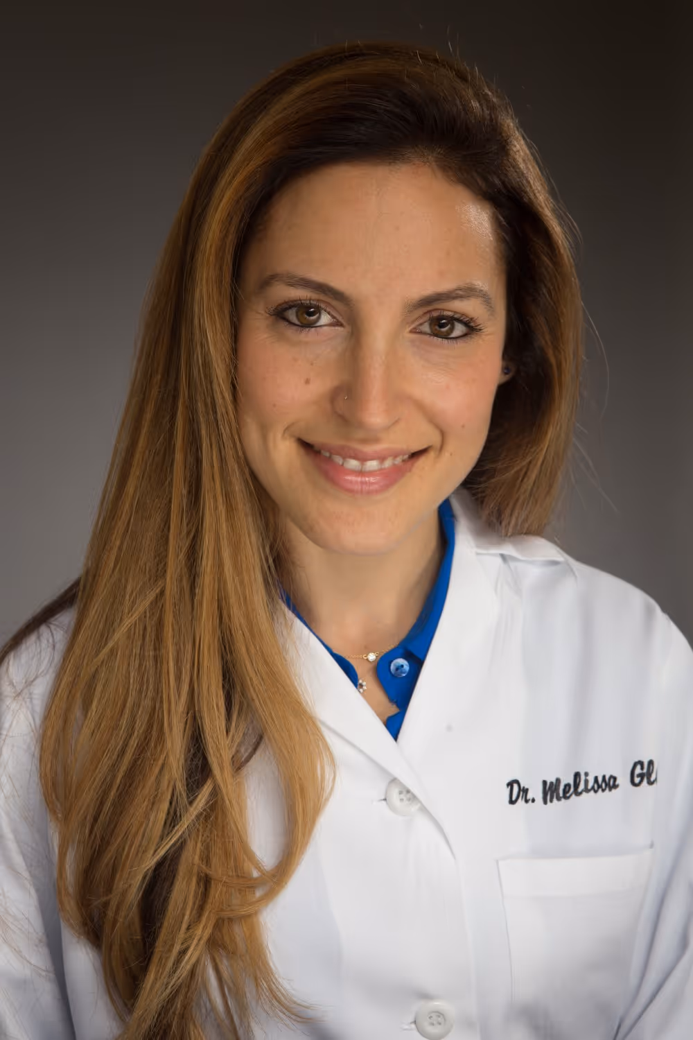 Smiling woman with long brown hair wearing a white lab coat and blue shirt against a gray background.