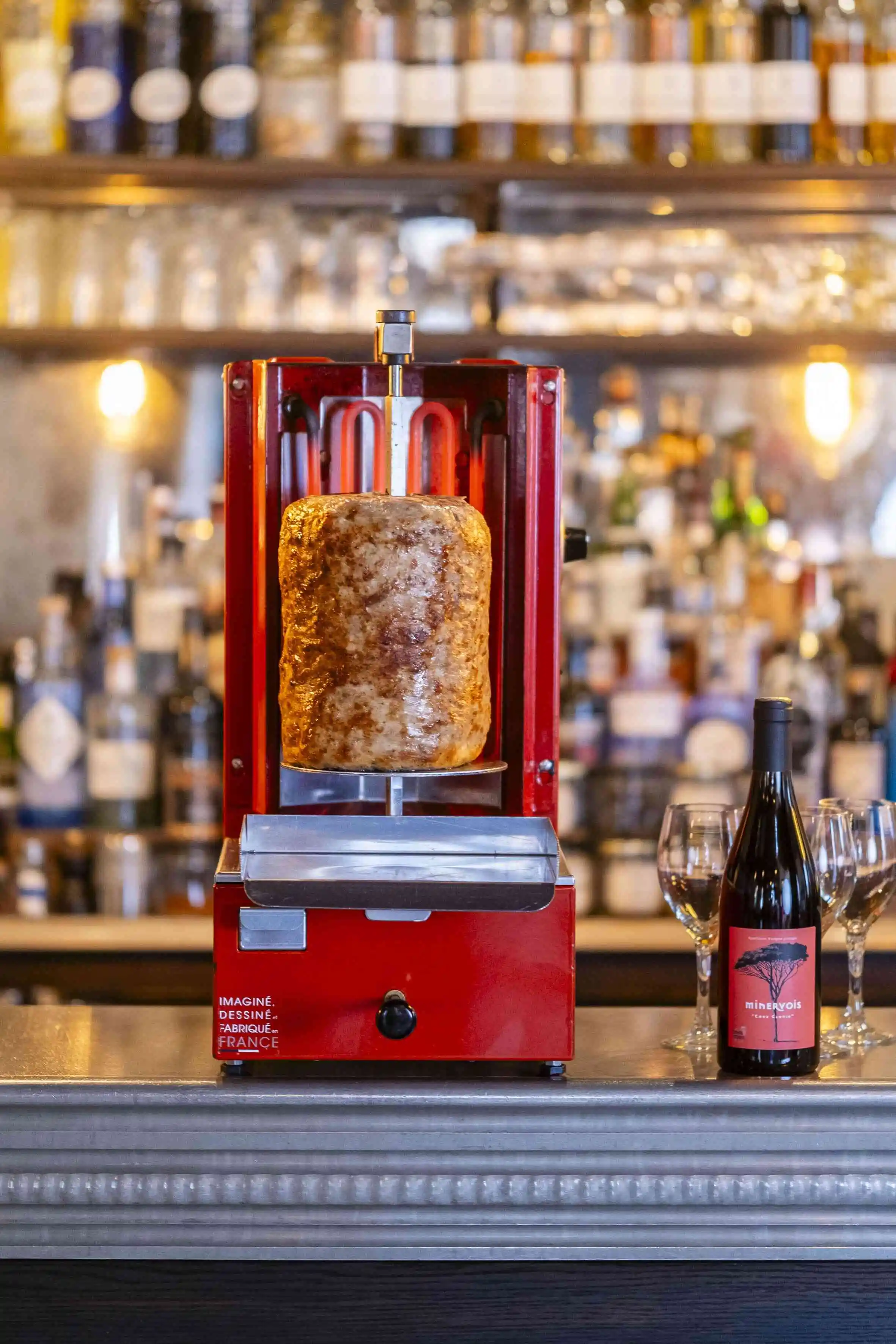 Rotisserie meat cooker with a large meat drum on the counter beside a wine bottle and glasses.