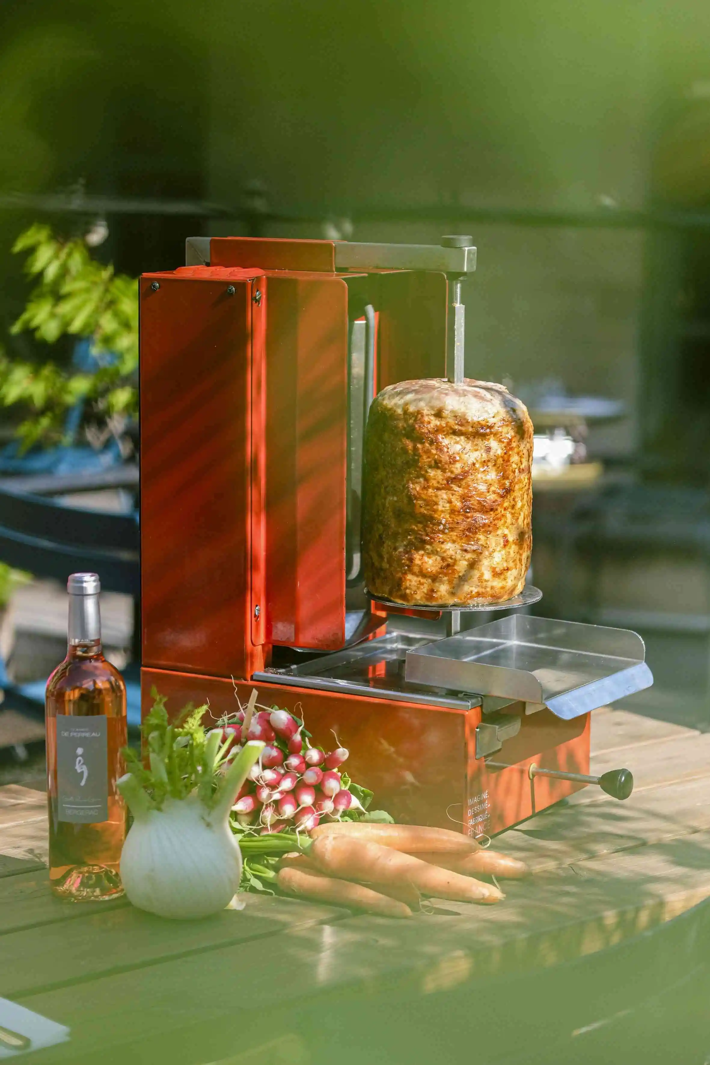 Red vertical rotisserie with cooked meat, carrots, radishes, fennel, and a bottle of rosé wine on wooden table.