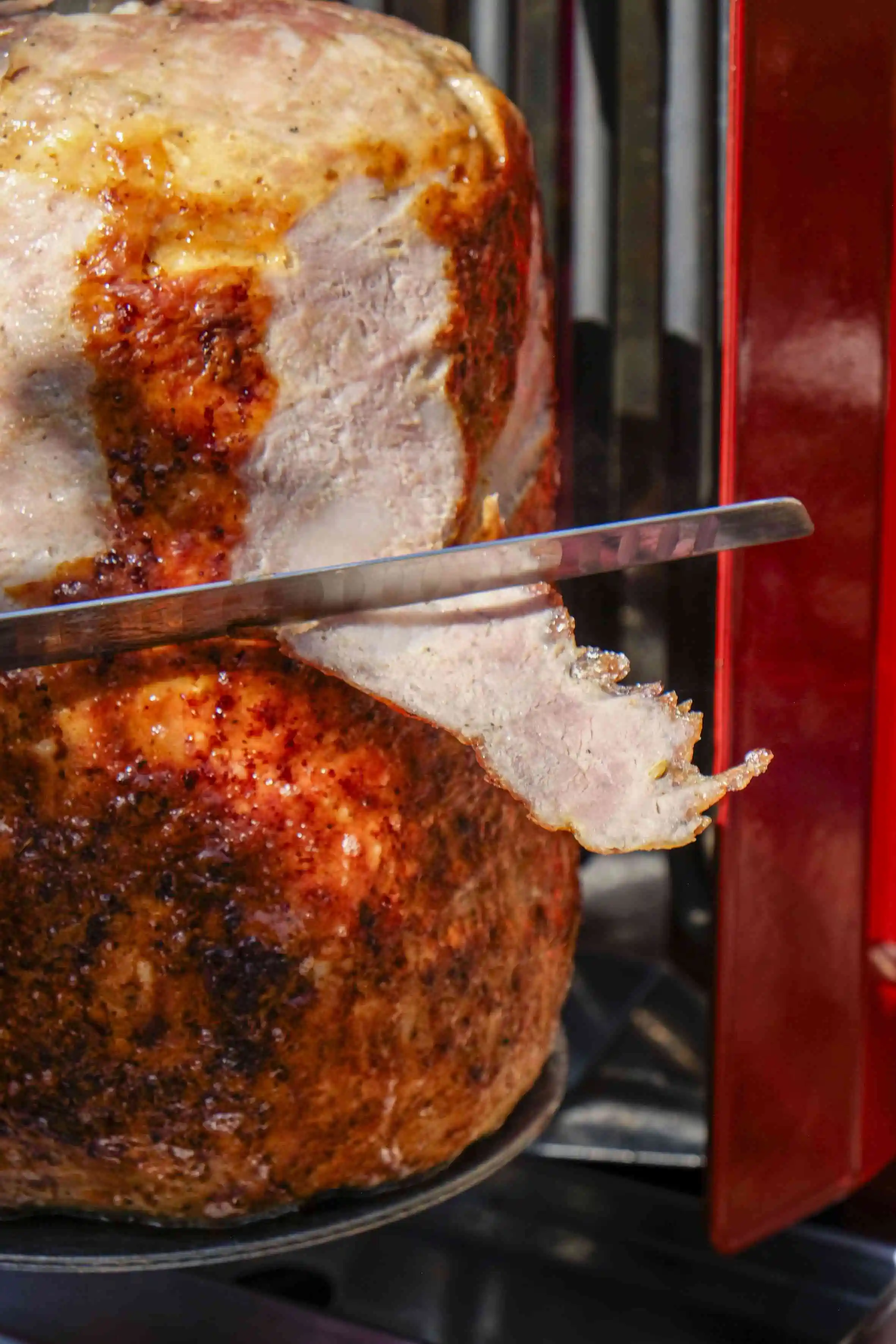 Slice of cooked, seasoned meat being cut with a long knife on a rotisserie spit.