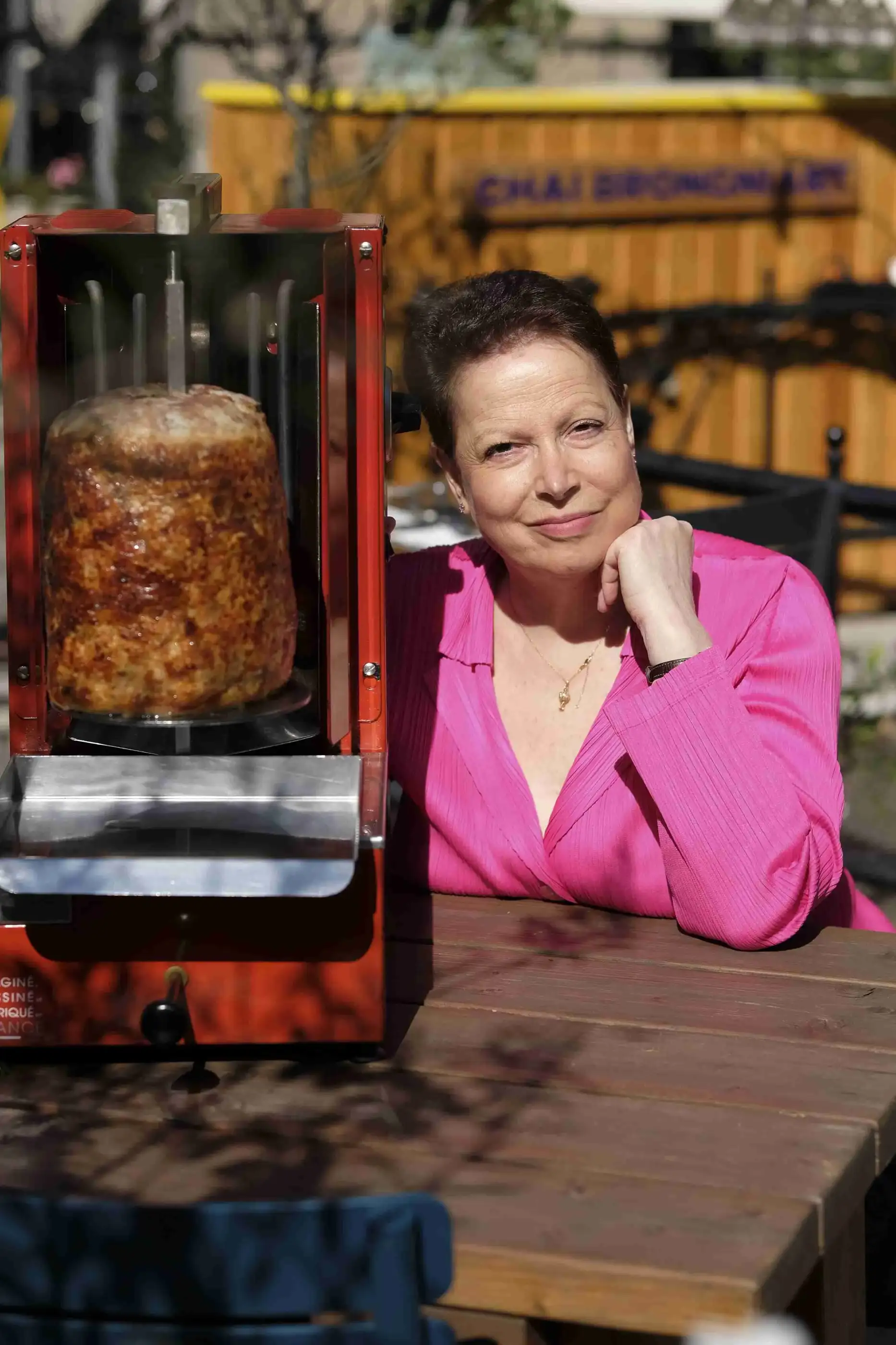 Woman in pink blouse sitting outdoors beside a vertical rotisserie with cooking meat.