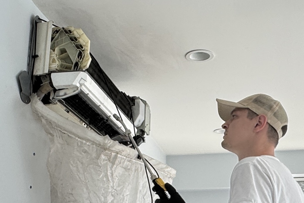 Technician wearing a cap and gloves repairing the internal components of a wall-mounted air conditioning unit.