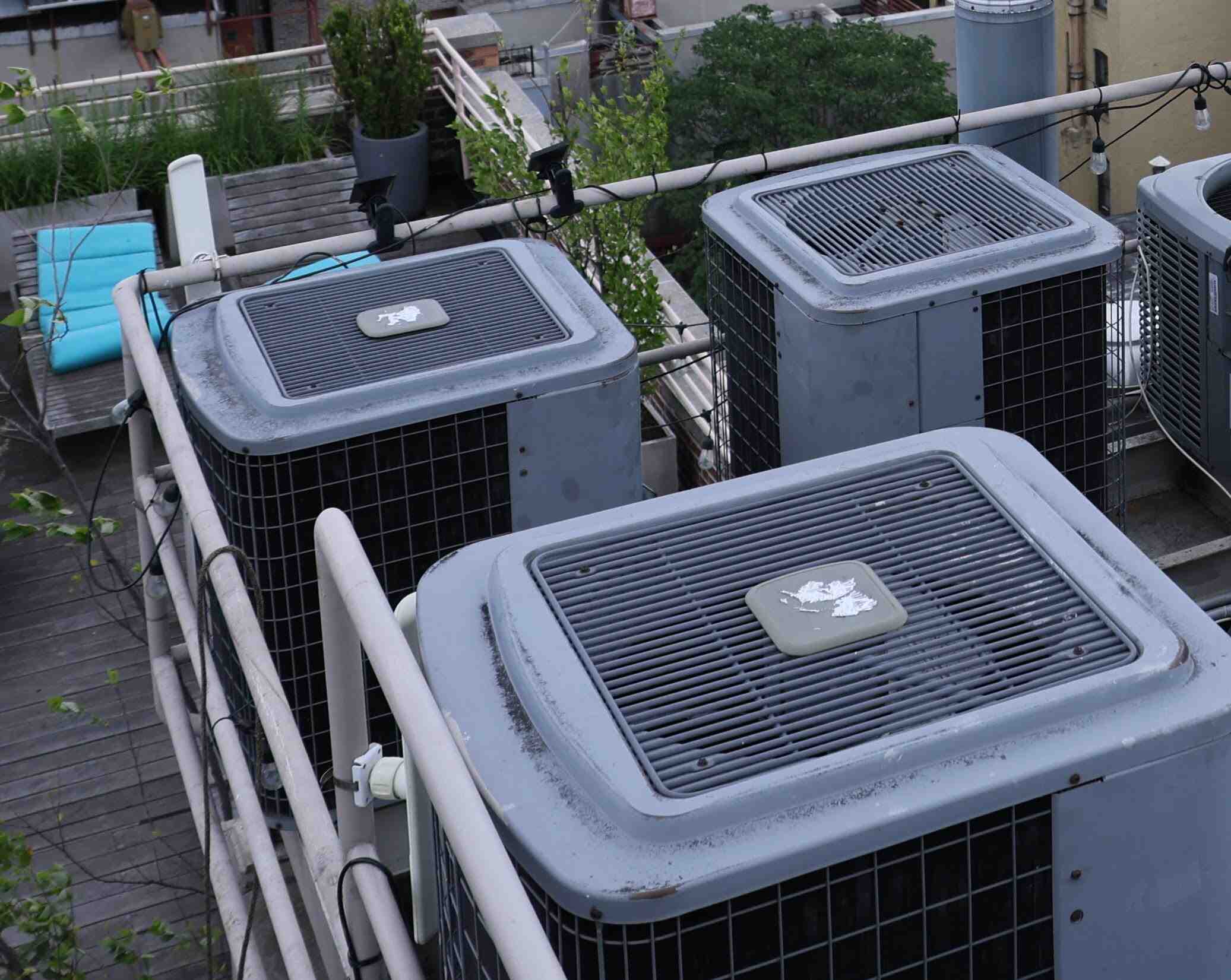 Close-up view of several rooftop air conditioning units with a metal railing and outdoor seating area in the background.