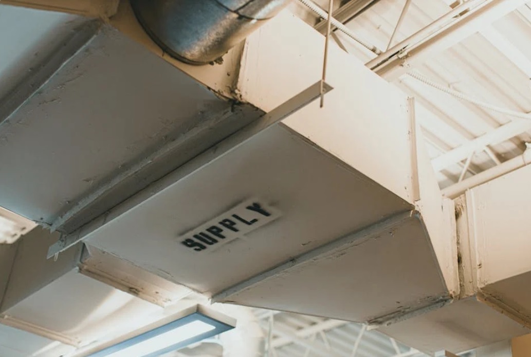 Metal air duct labeled 'SUPPLY' suspended from a ceiling with exposed pipes and lighting.