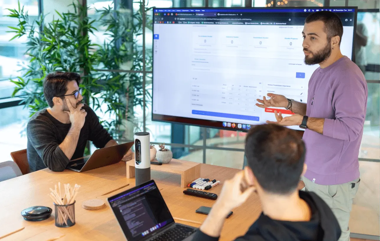Team members gathered around a screen as one presents project data during a meeting in the office.