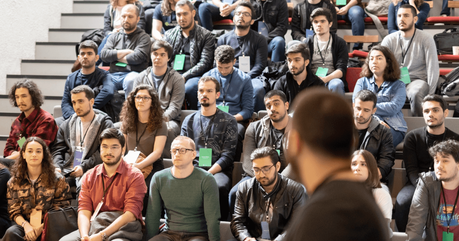 A speaker stands on stage addressing a large, seated audience in a university auditorium during a tech or professional event.