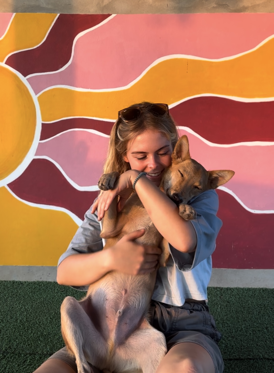 Smiling young woman sitting on grass holding and hugging a relaxed dog against a colorful mural with sun rays.