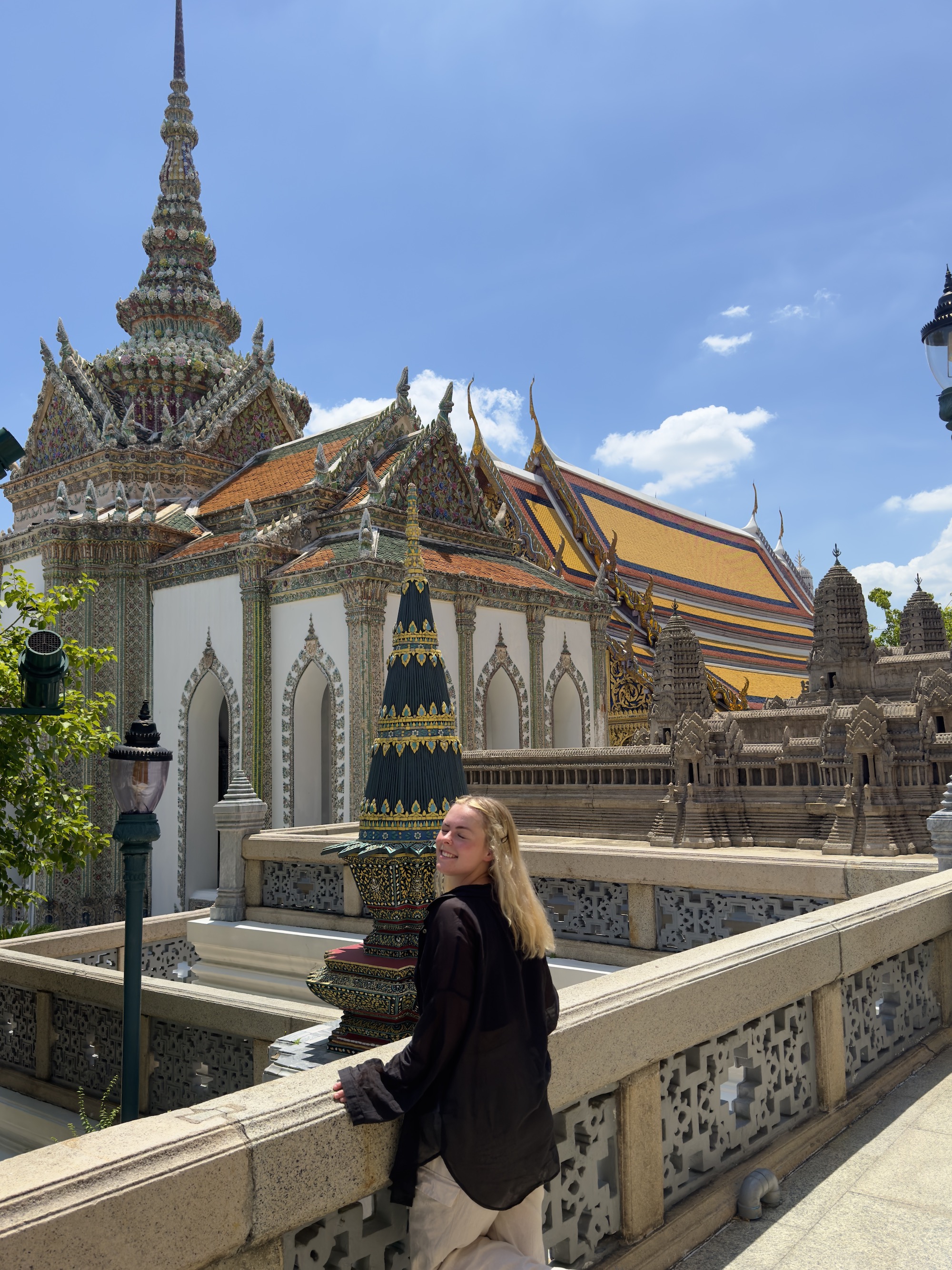 Smiling woman in black shirt leaning on stone railing near ornate temple buildings under a clear blue sky.