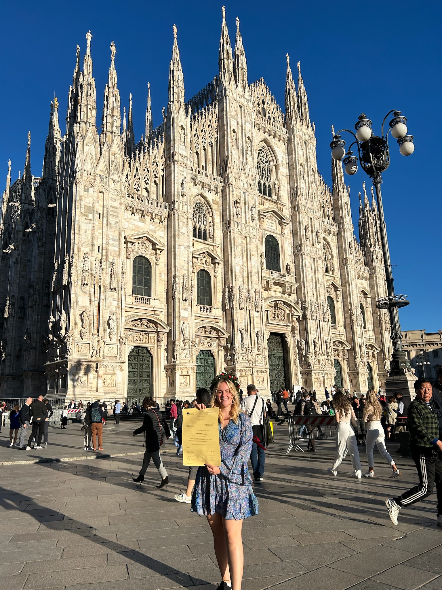 Young woman smiling and holding a large yellow certificate, standing in front of the ornate marble facade of Milan Cathedral under a clear blue sky.