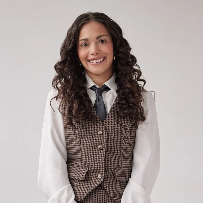 Smiling woman with long curly dark hair wearing a white shirt, dark tie, and brown houndstooth vest against a light background.