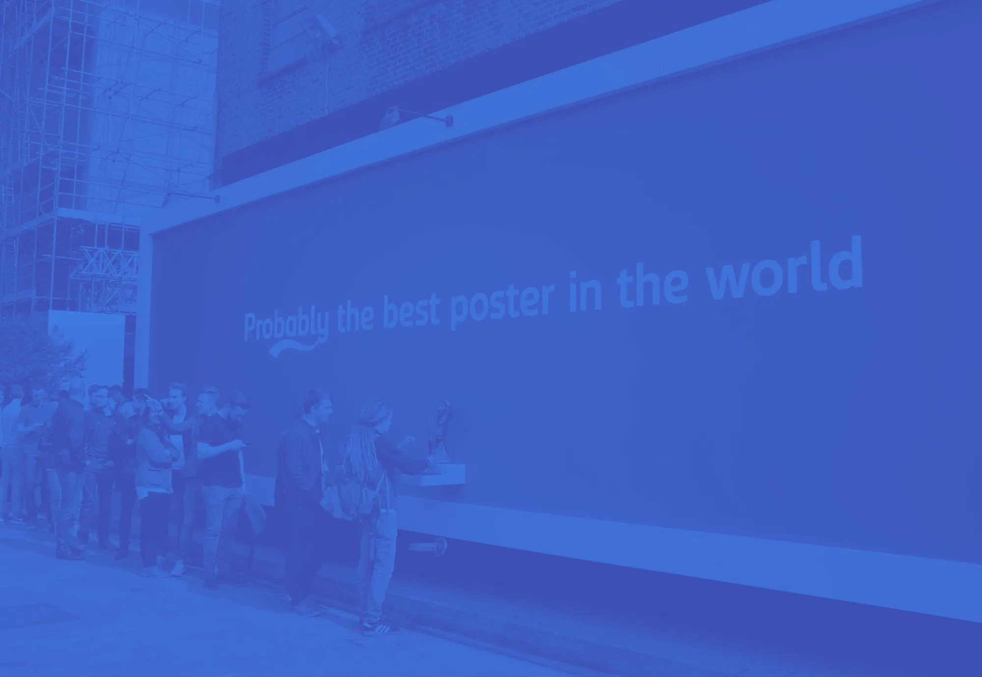 People standing in line outside near a wall with the text 'Probably the best poster in the world.'