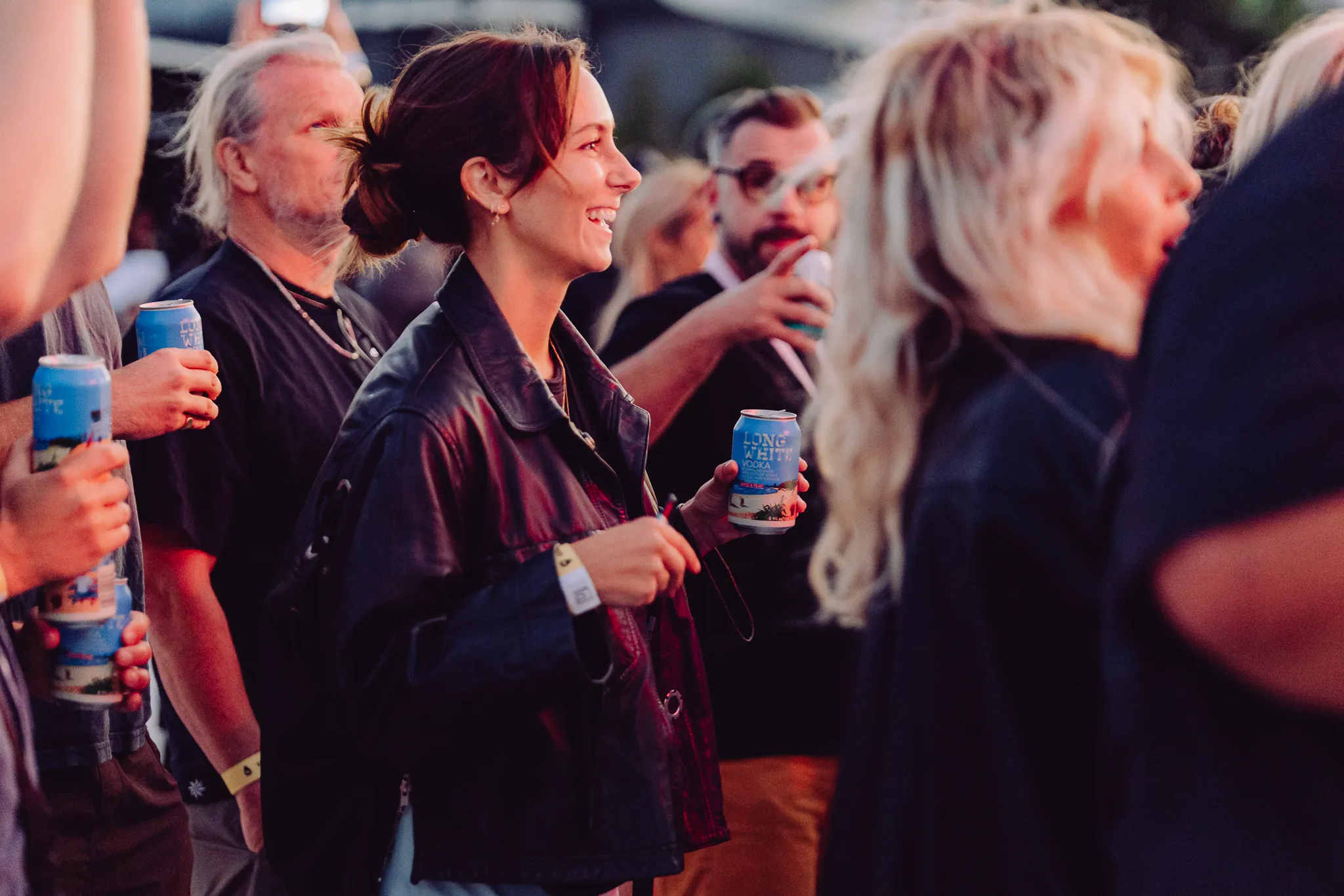 Group of people at an outdoor event holding cans of Long White Vodka, with a smiling woman in a leather jacket.
