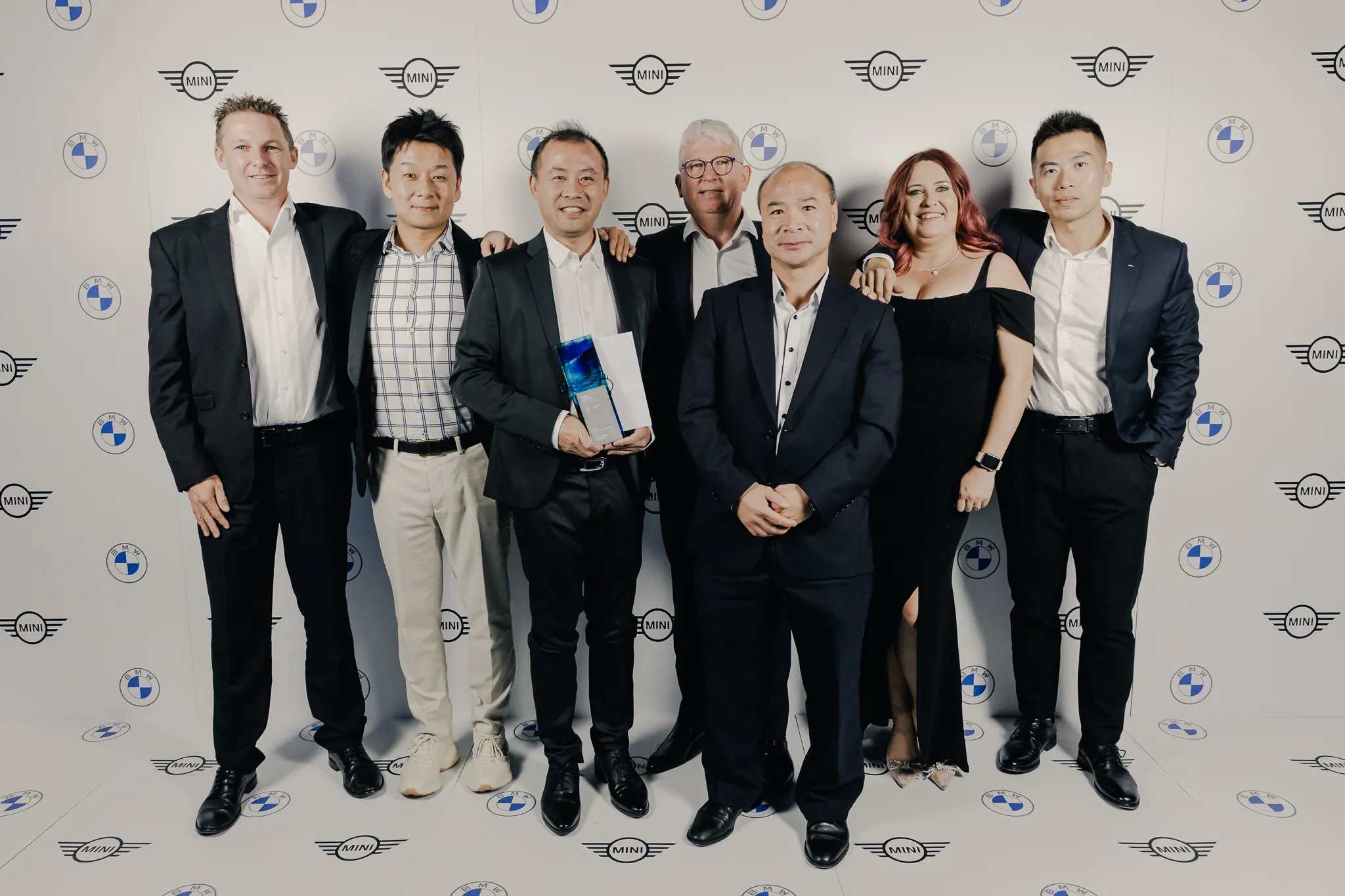 Seven people in formal attire posing at a BMW and Mini event, one holding an award.