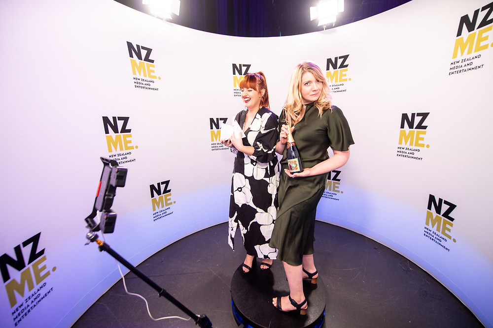 Two women posing on a small round platform with NZME media backdrop and a camera on a tripod.