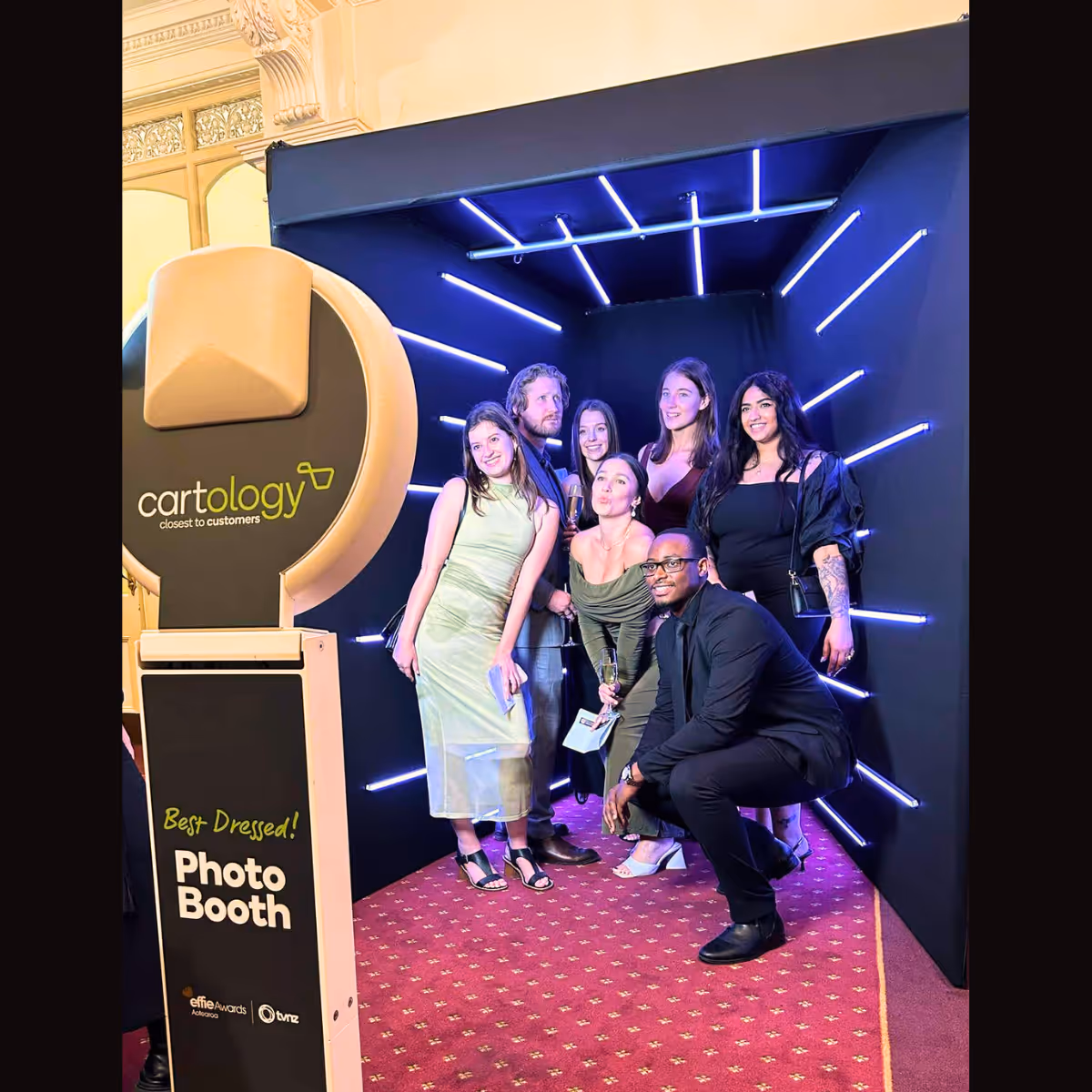 Group of six people posing inside a photo booth with neon lights and red carpet at an event.
