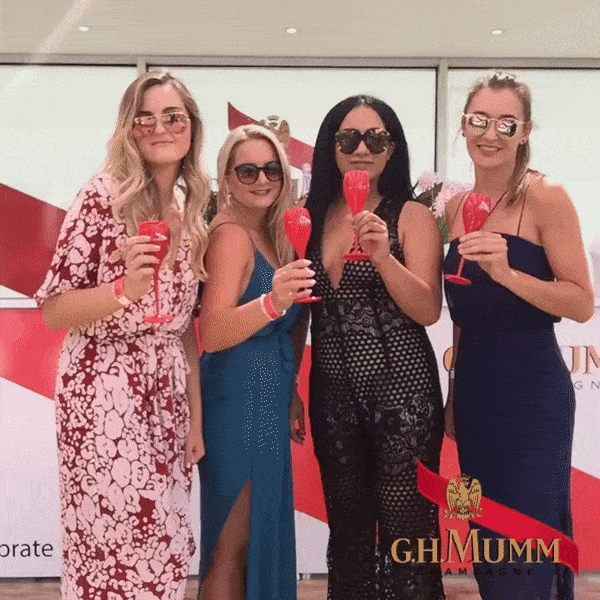 Four women in dresses and sunglasses holding red glasses, posing and smiling at a G.H. Mumm event.