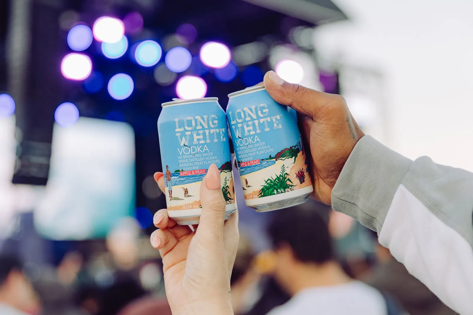 Two hands clinking cans of Long White vodka sparkling water with apple and pear flavor at an outdoor event.