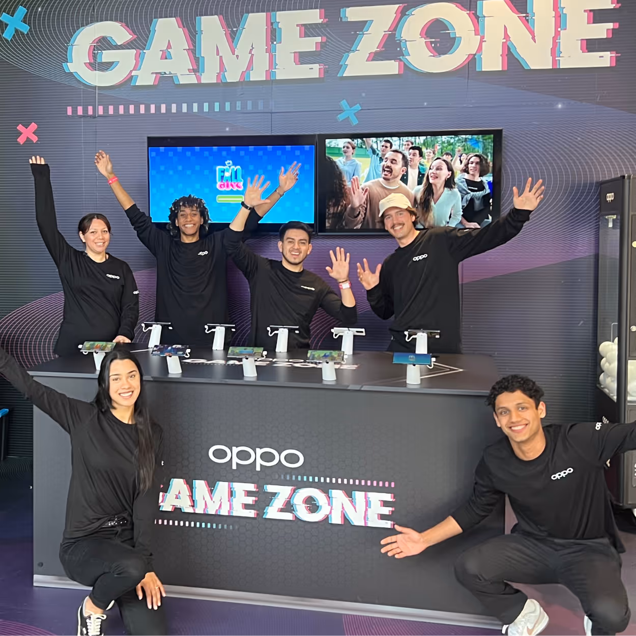 Six people in black Oppo shirts smiling and raising hands behind a game zone booth with mobile devices.