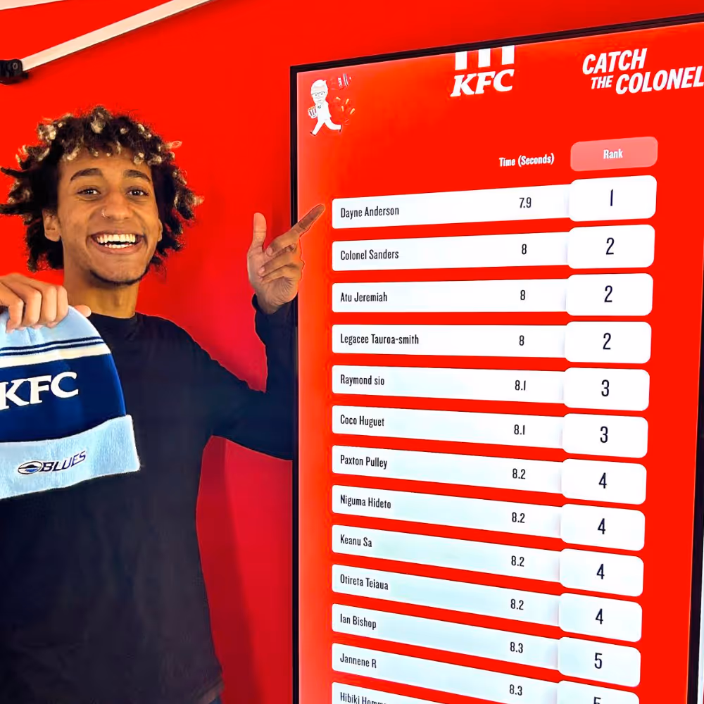 Smiling man in black points to KFC leaderboard showing names, times in seconds, and ranks against red wall.