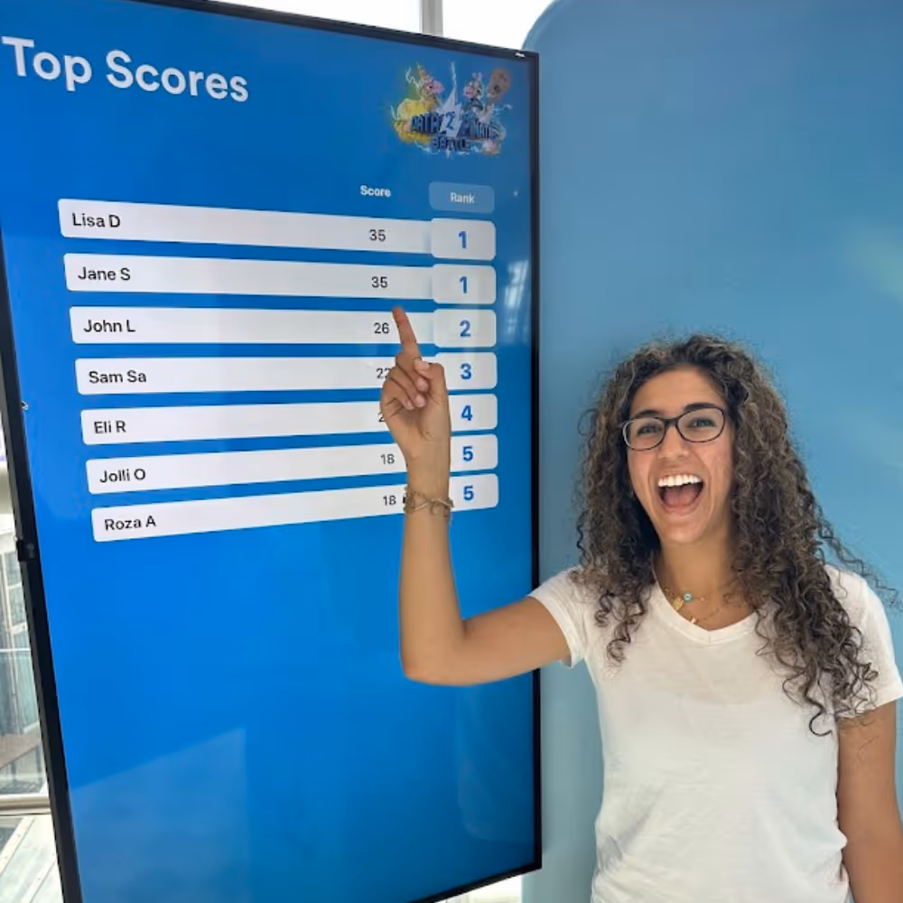 Smiling woman with glasses points to tied top score on a large blue leaderboard screen.