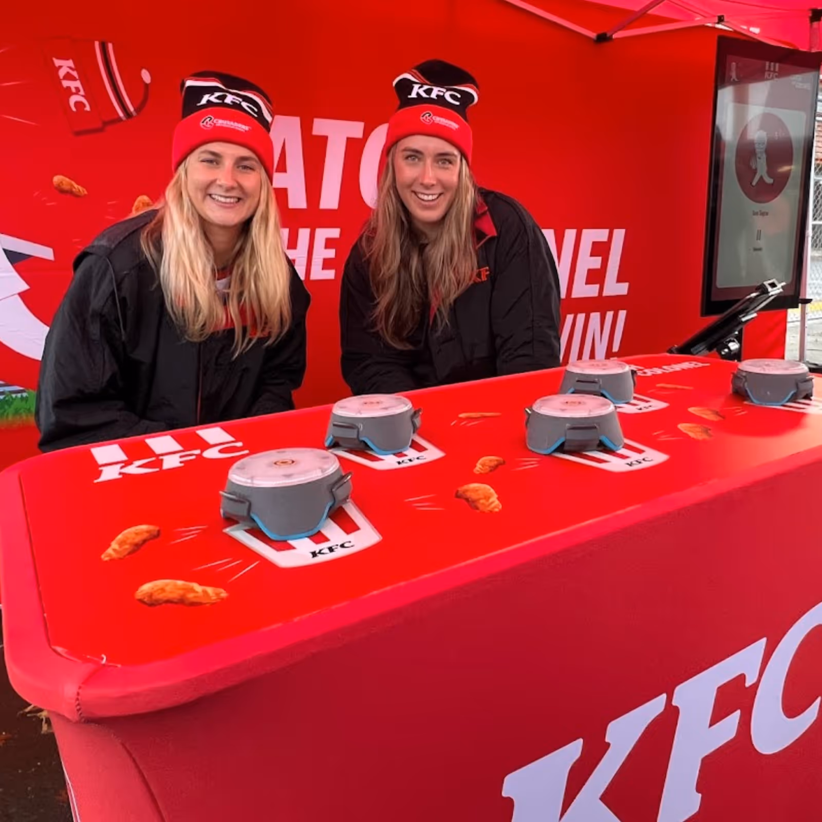 Two women in KFC beanies and jackets smiling behind a red KFC branded table with six buzzer buttons.