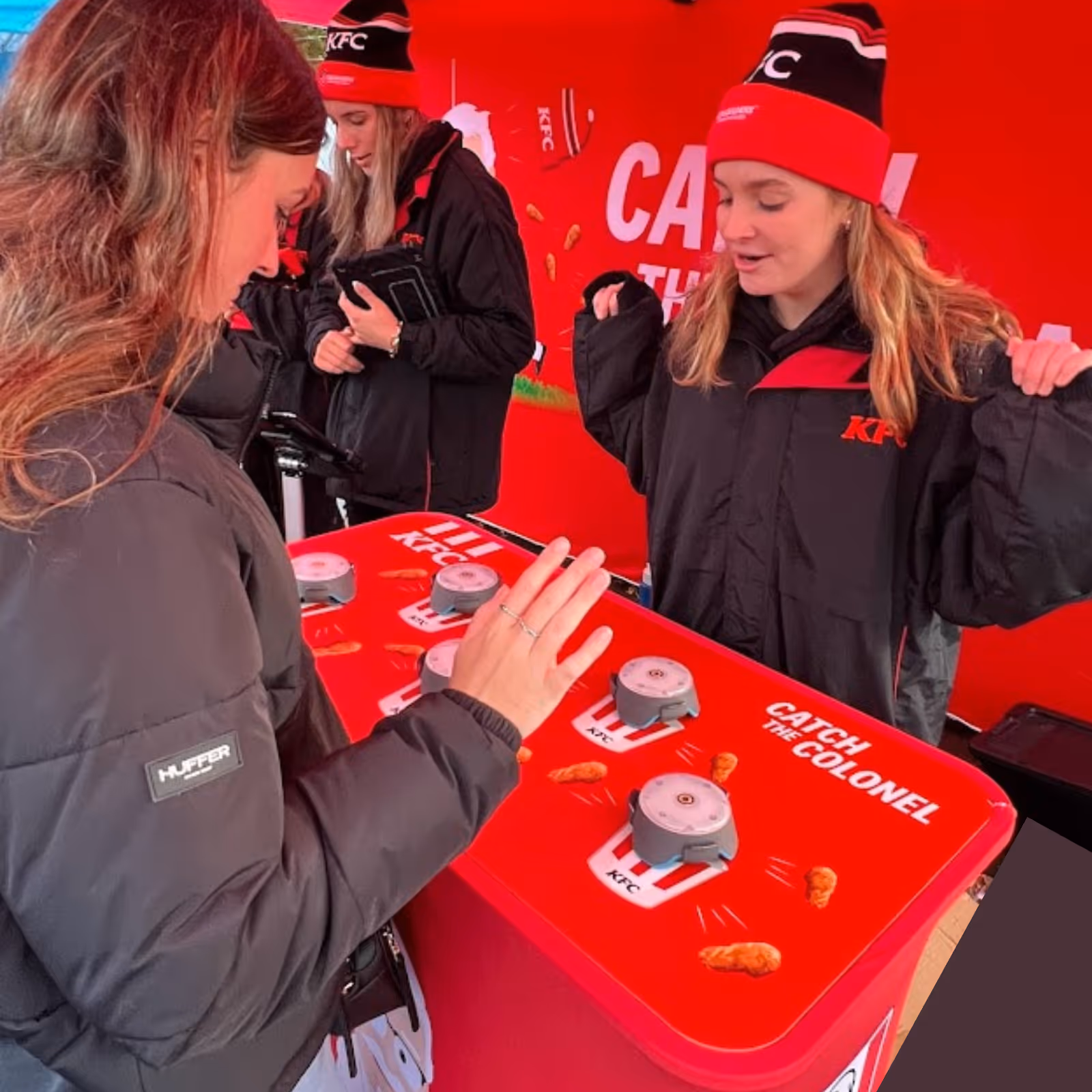 Woman playing KFC's Catch the Colonel game at a red promotional booth with two staff in black KFC jackets.