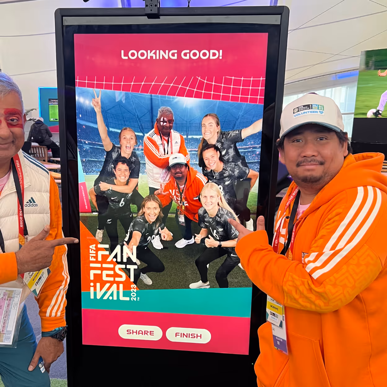Two men in orange jackets pointing at a digital screen with a photo of a group of soccer fans.