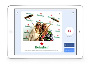Two women posing with photo booth props in front of a Heineken branded backdrop on a tablet screen.
