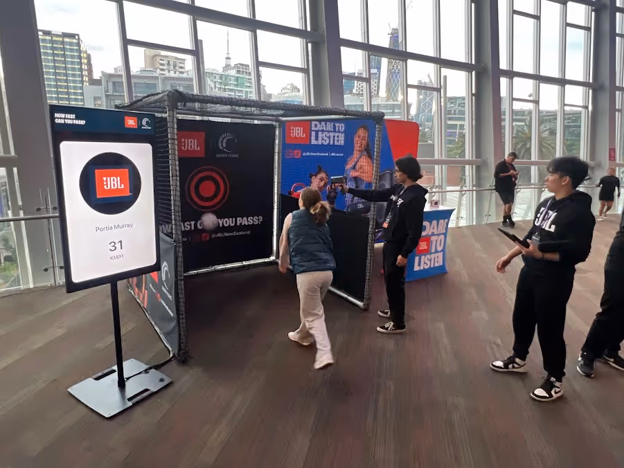 Indoor event where a woman throws a ball at a speed target, tracked by people with JBL gear.