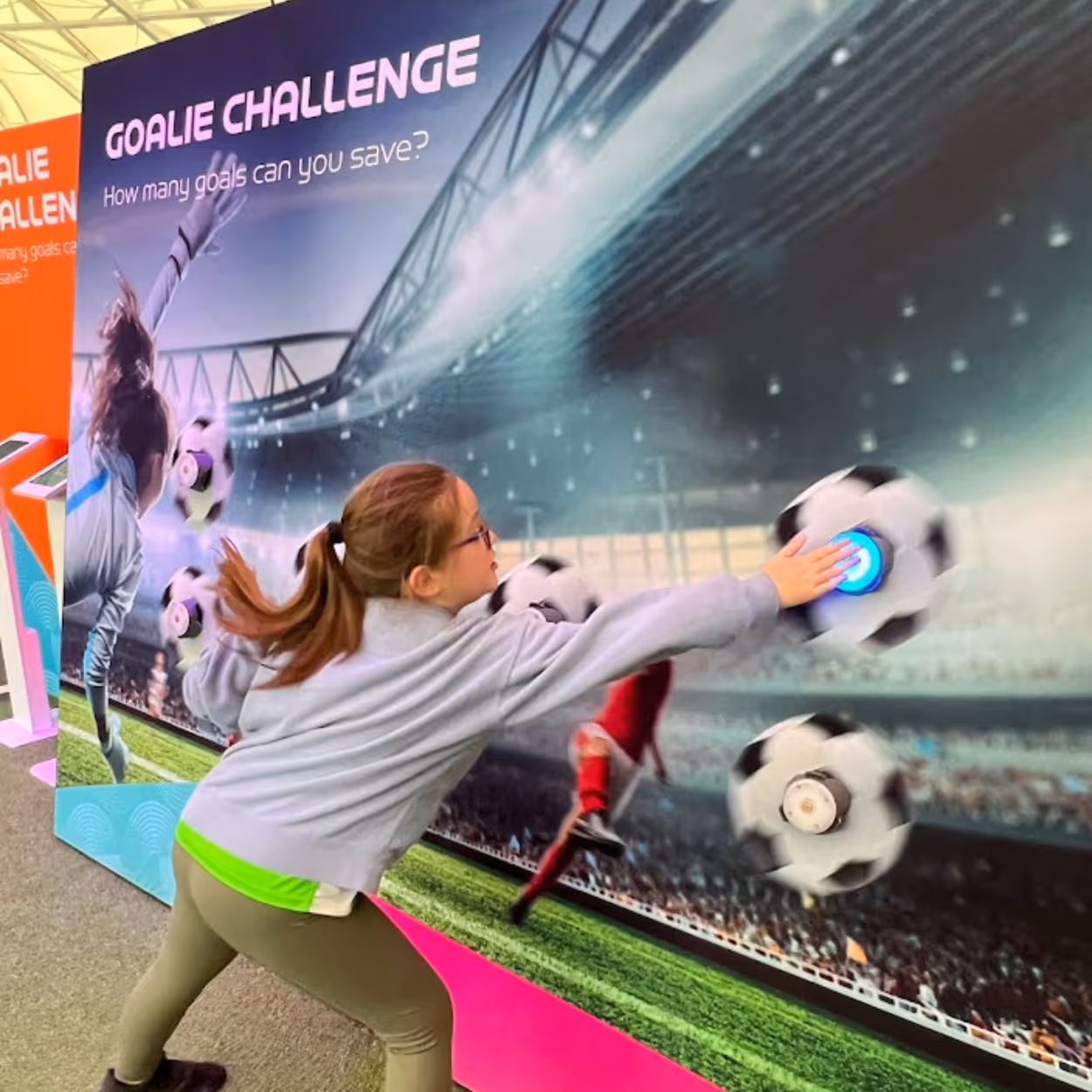 Girl reaching to touch an interactive soccer ball on a wall in a 'Goalie Challenge' game display.