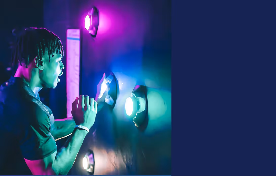 Man with dreadlocks playing an illuminated reaction game with glowing buttons on a dark wall.