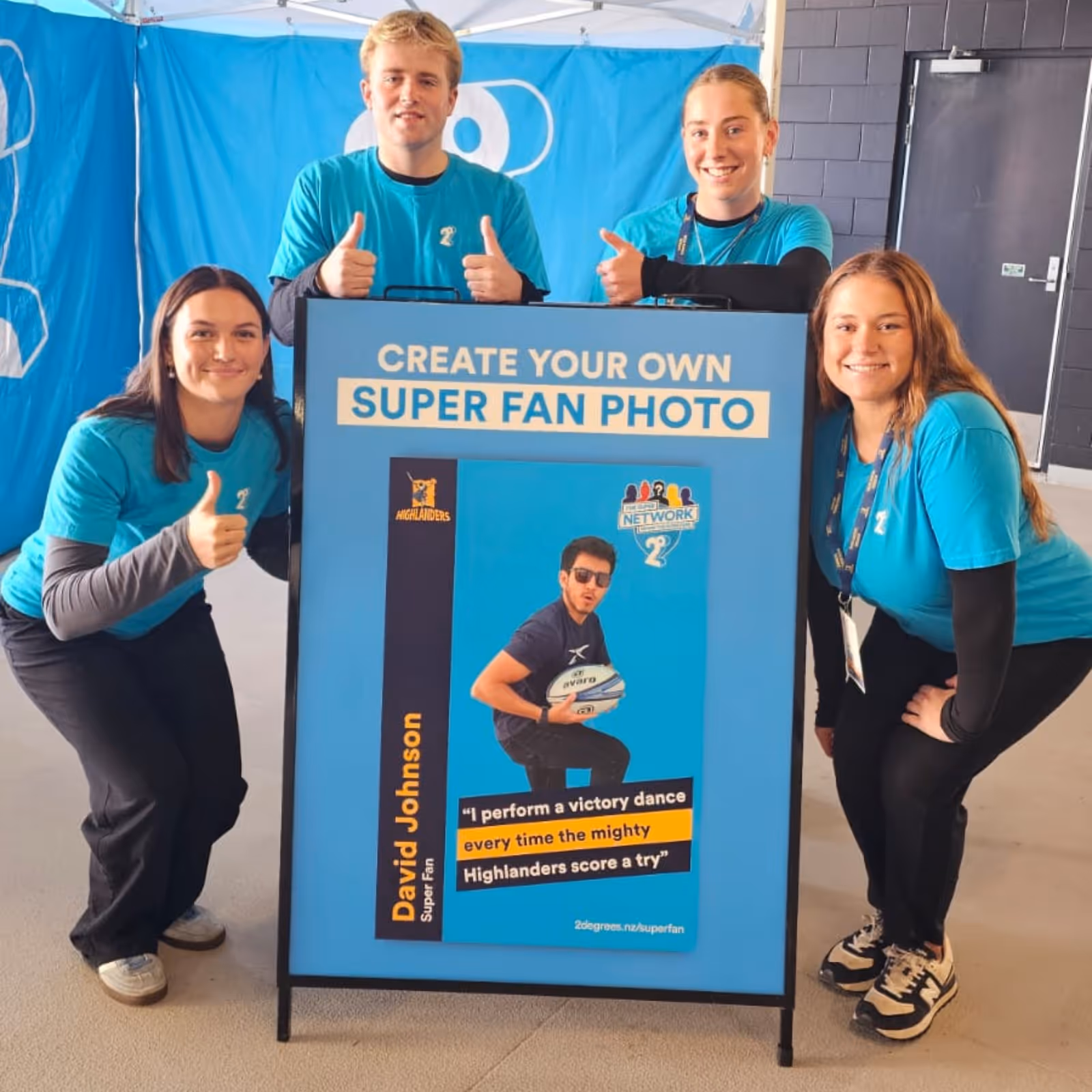 Four people in blue shirts giving thumbs up around a sign promoting super fan photos with a rugby theme.
