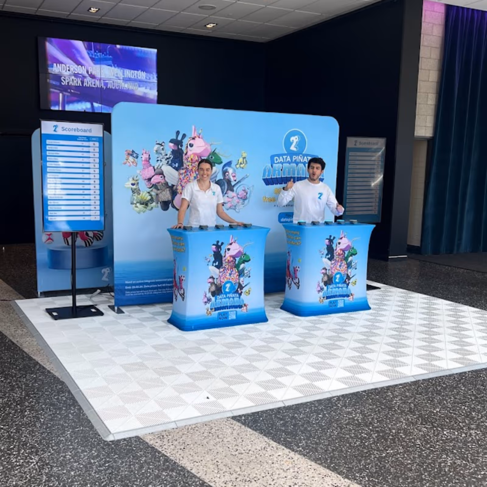 Two people at bright blue promotional booths with mascot images and scoreboards behind in event space.