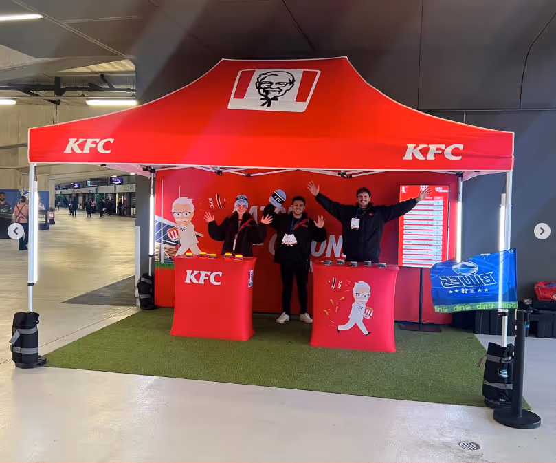 KFC promotional booth with red canopy, two counters, and three staff waving indoors at an event.